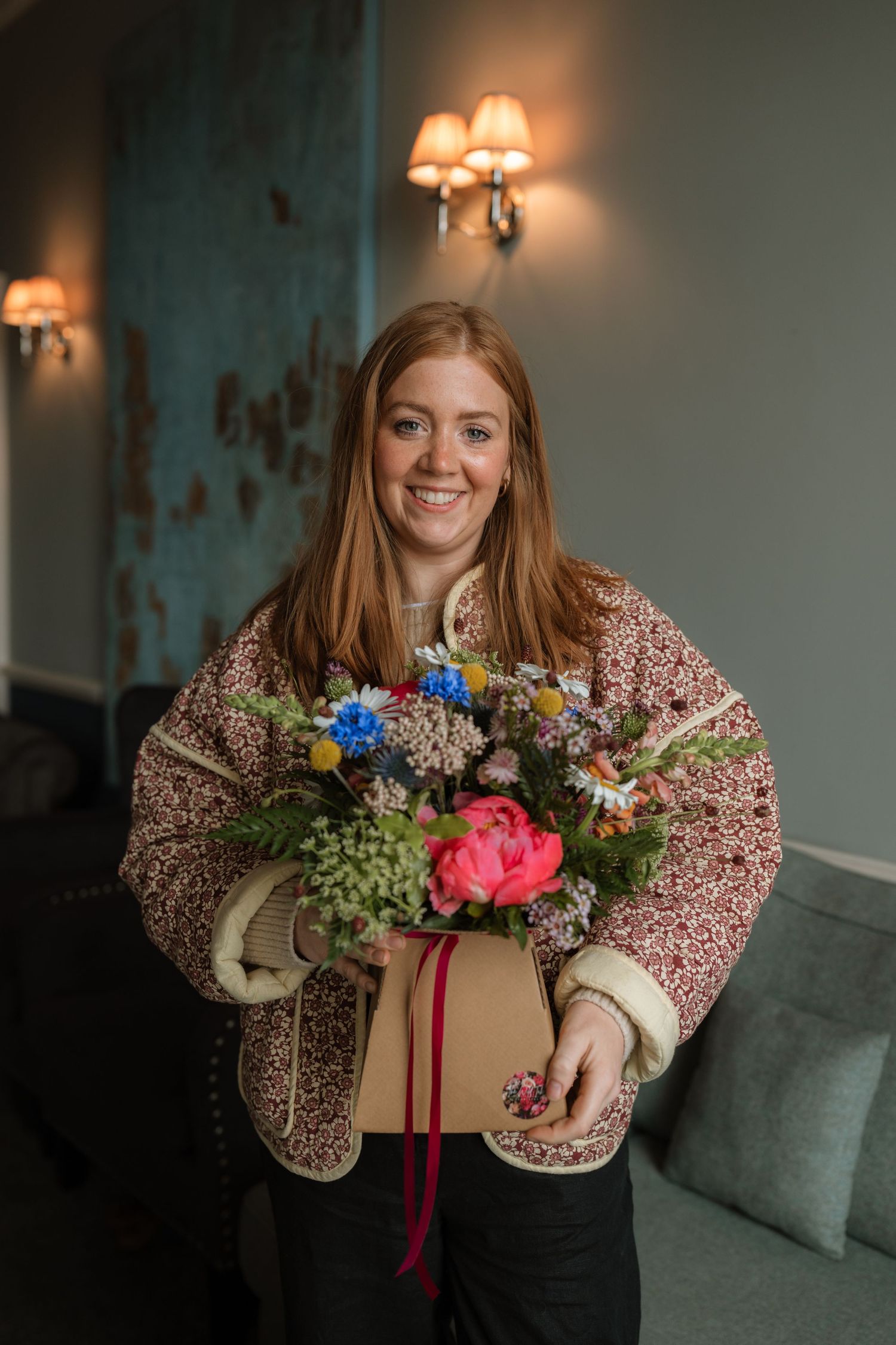 A person in a floral print jacket holds a beautiful arrangement of colorful flowers with red ribbon in a cozy room.