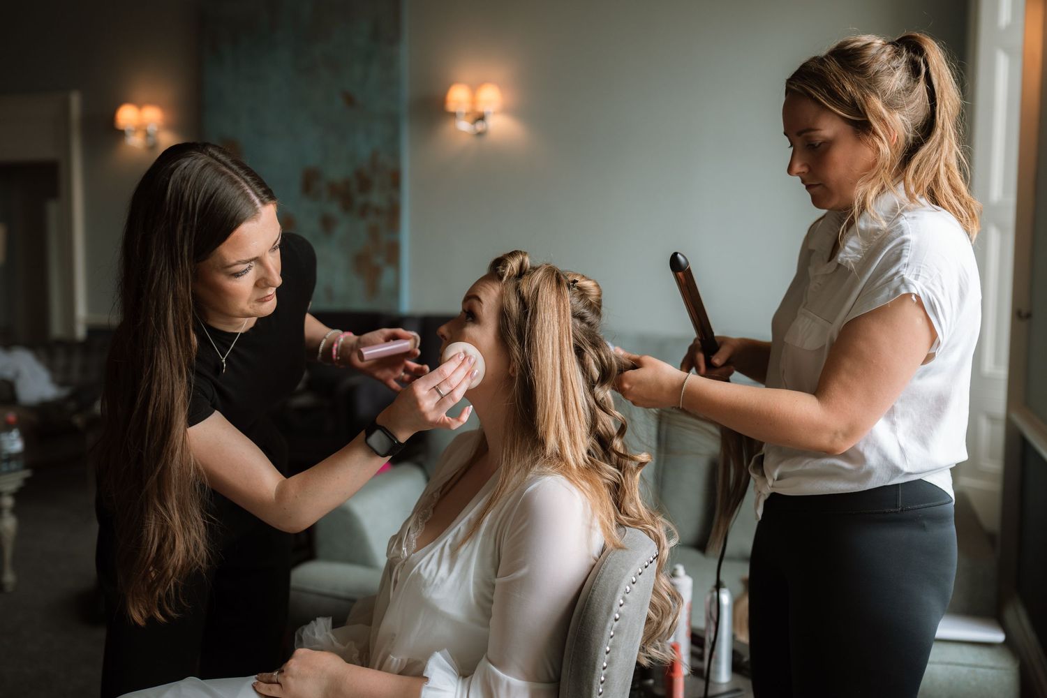 Hair stylists work together on bridal preparation, styling hair in an elegant room with wall sconces.