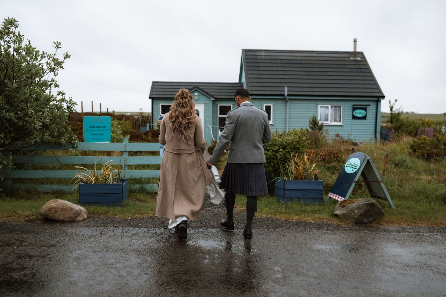 A couple walking together in the rain towards a small blue cottage with a turquoise fence in a rural setting.