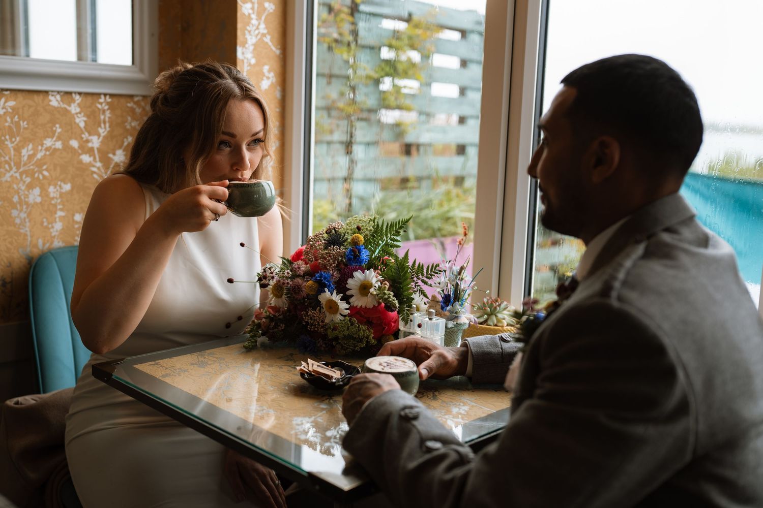 A couple sharing wine and conversation at a restaurant table near a window with a colorful flower arrangement.