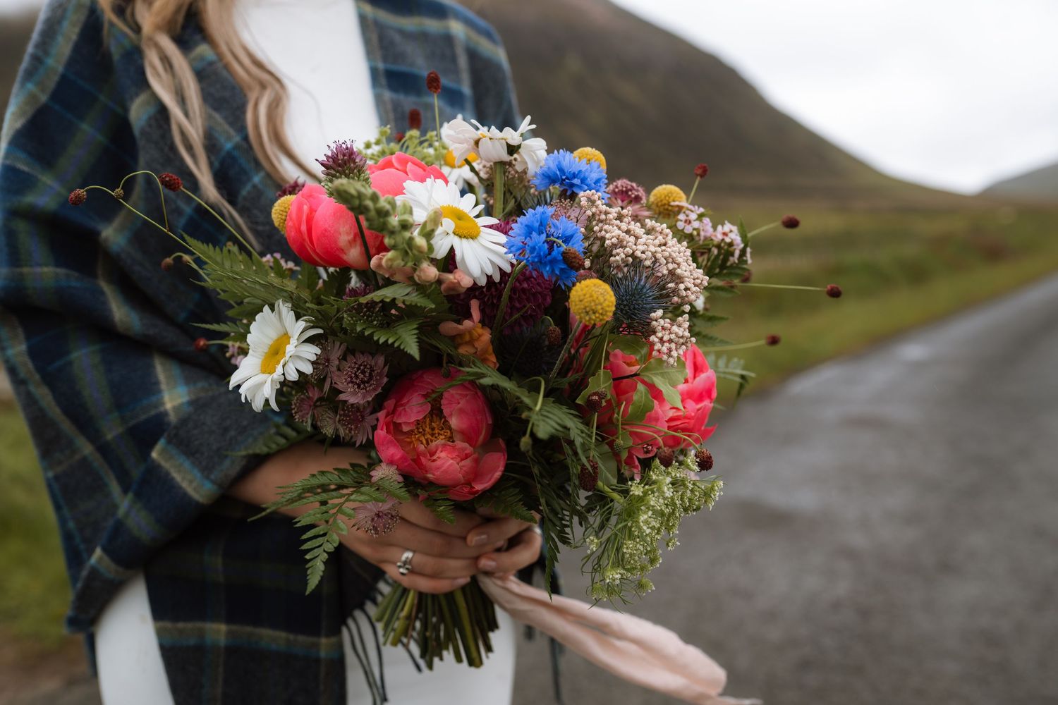 Vibrant wedding bouquet with pink peonies, blue cornflowers, daisies and mixed wildflowers against highland scenery.