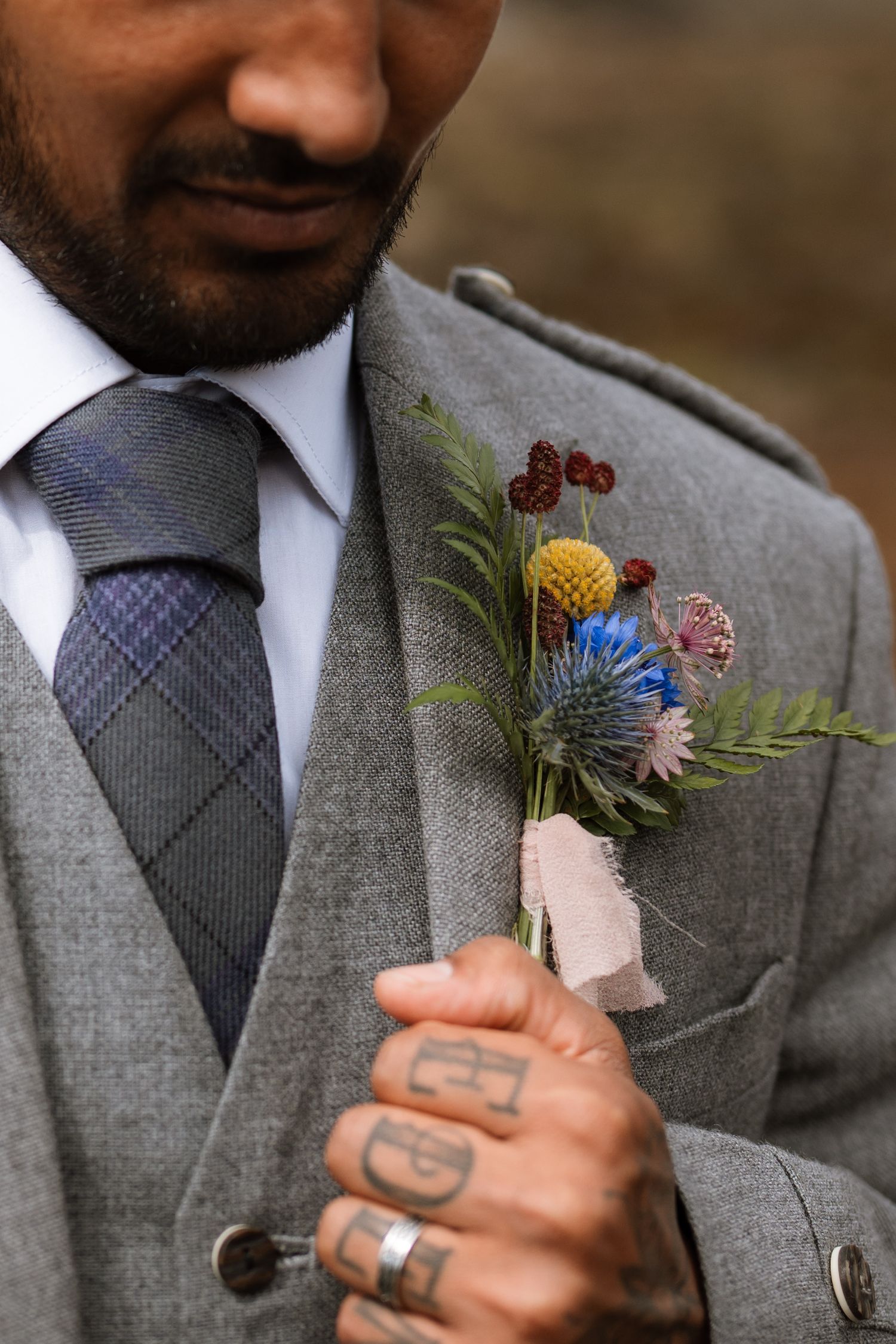Close up of colorful wildflower boutonniere pinned to grey suit lapel with tattooed hands visible.