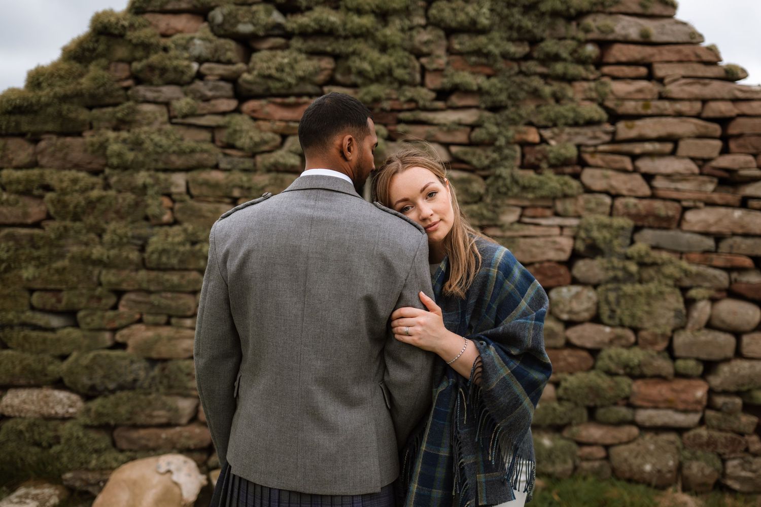 Two people share an intimate embrace against a rustic stone wall backdrop during a romantic outdoor photoshoot.