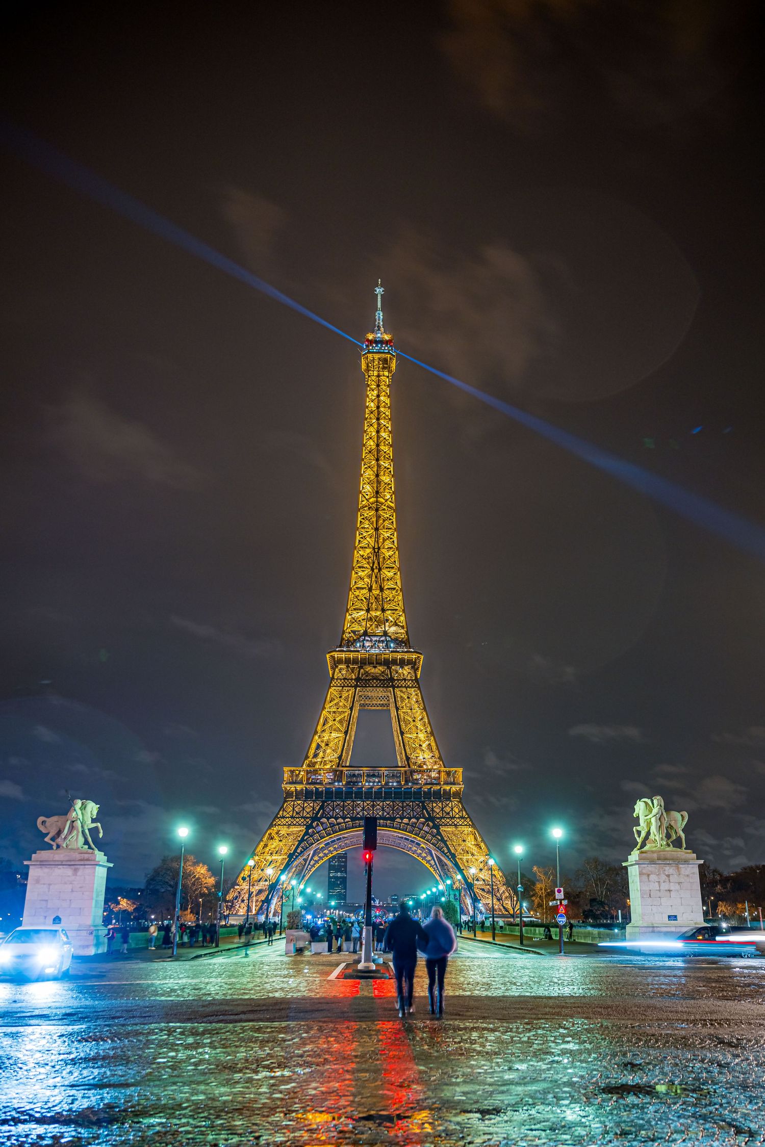 Illuminated Eiffel Tower glows golden against dark night sky with wet reflective pavement and blue light beams above.