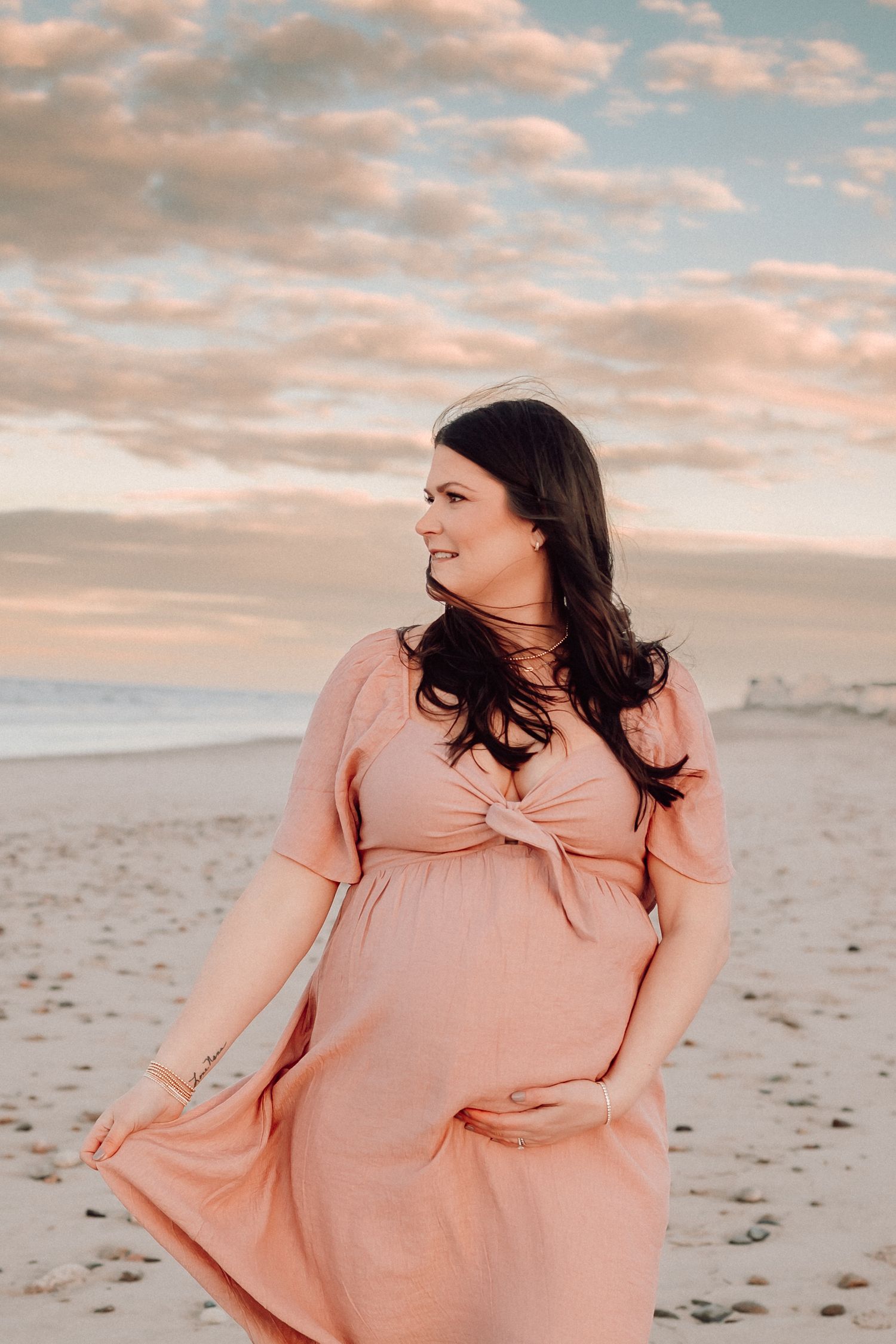 Woman in flowing peach dress stands on beach during sunset with clouds reflecting pink hues.