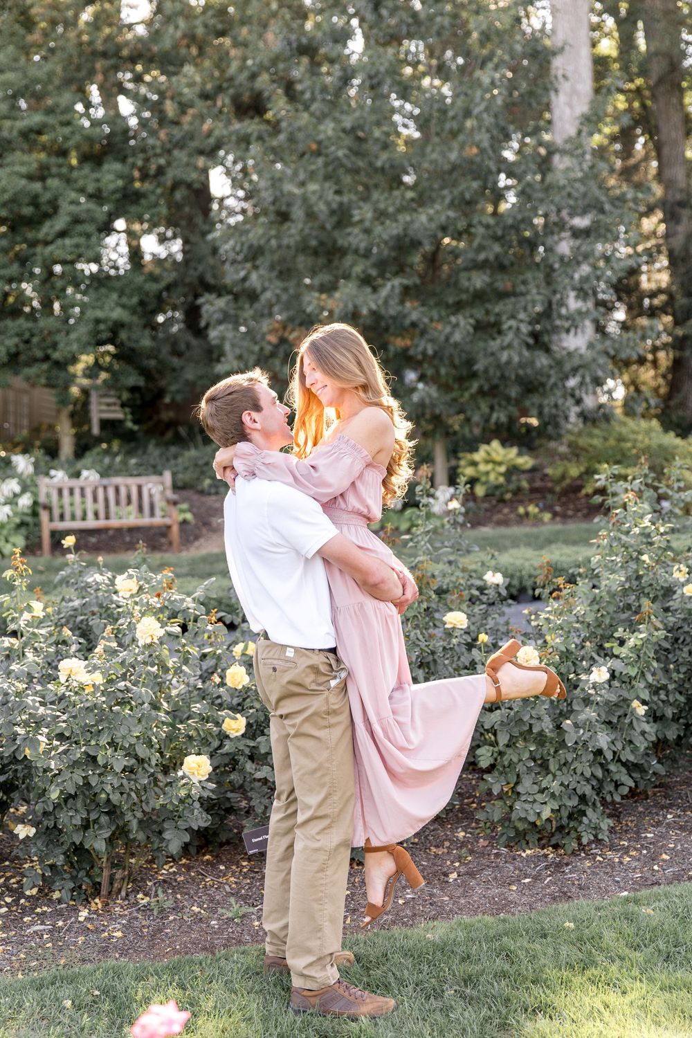 Romantic couple embraces in garden setting with blooming roses and greenery at sunset.