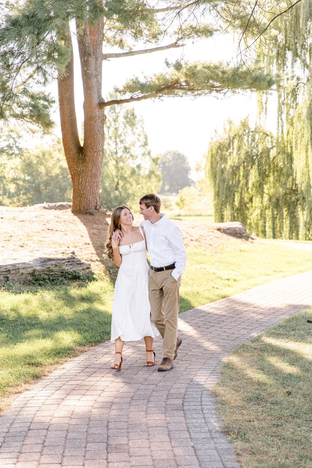 A couple walks together on a brick path through a sunlit park with willow trees and greenery in the background.