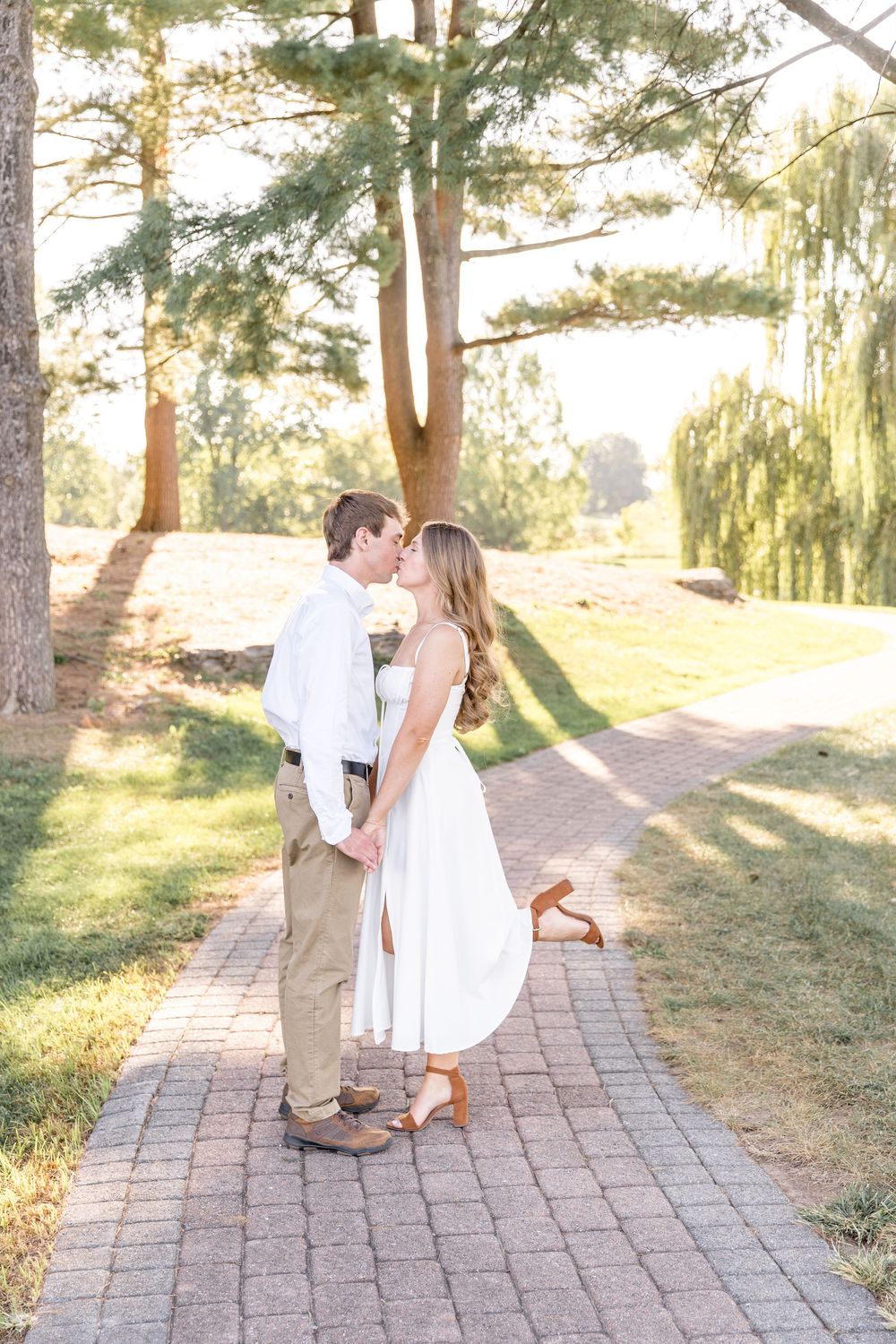 A couple shares a romantic moment on a brick path surrounded by tall pine trees and sunlit greenery at sunset.
