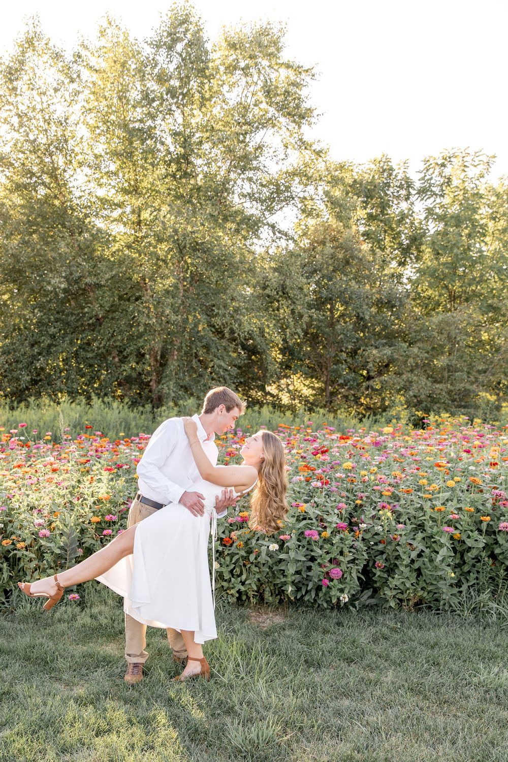 A couple in white outfits share a romantic dip pose in front of a blooming flower garden at sunset.