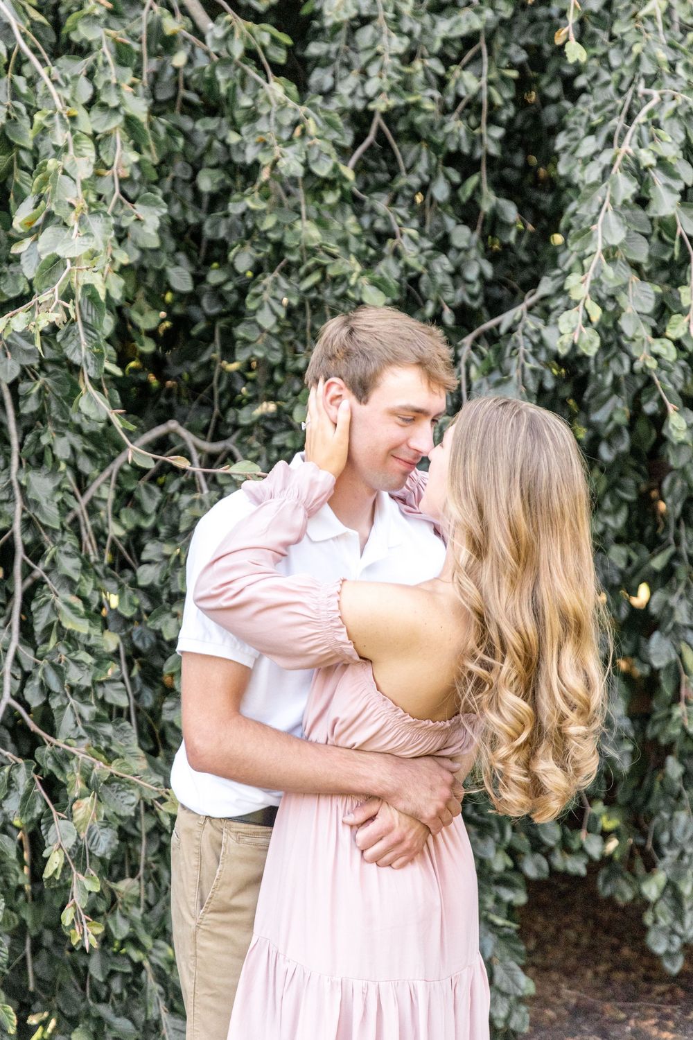Romantic couple embraces outdoors against lush green foliage backdrop, she in pink dress and he in white shirt.