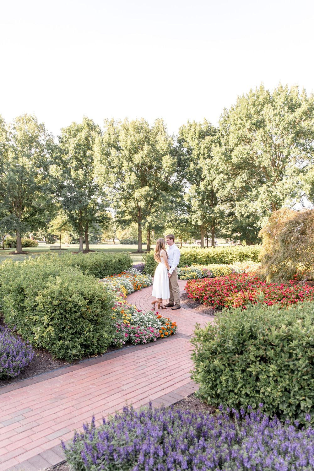 Two people embrace on a brick pathway surrounded by vibrant flower gardens and tall trees on a sunny day.