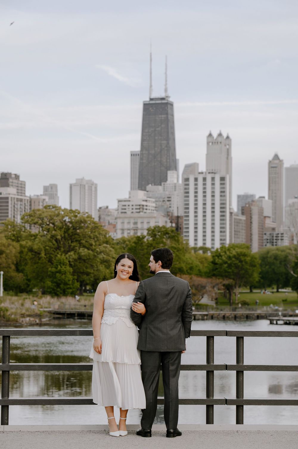 A couple stands on a pier overlooking the Chicago skyline with iconic John Hancock Building on an overcast day.
