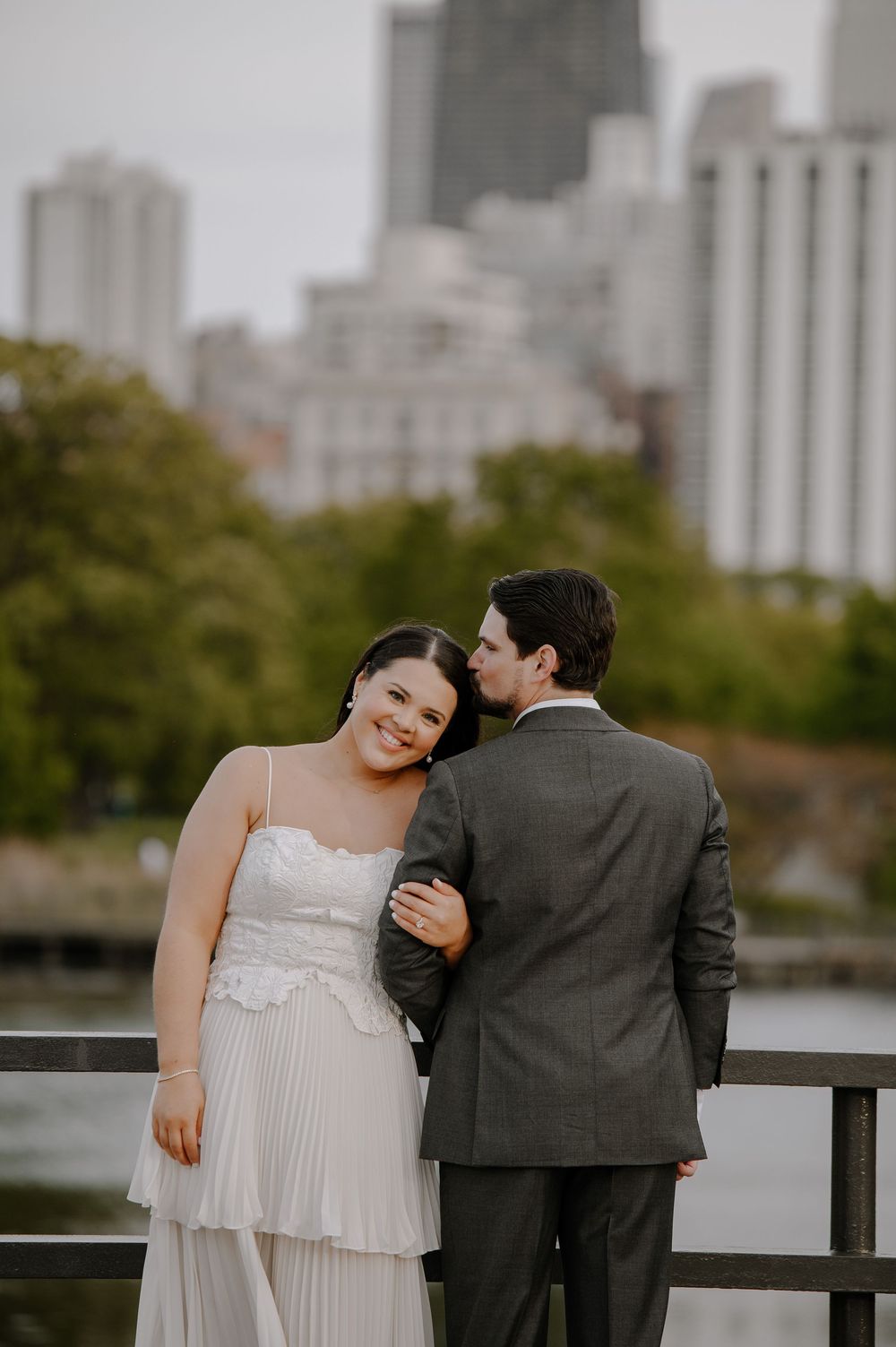 A couple in wedding attire share an intimate moment on a city rooftop with urban buildings and trees in the background.