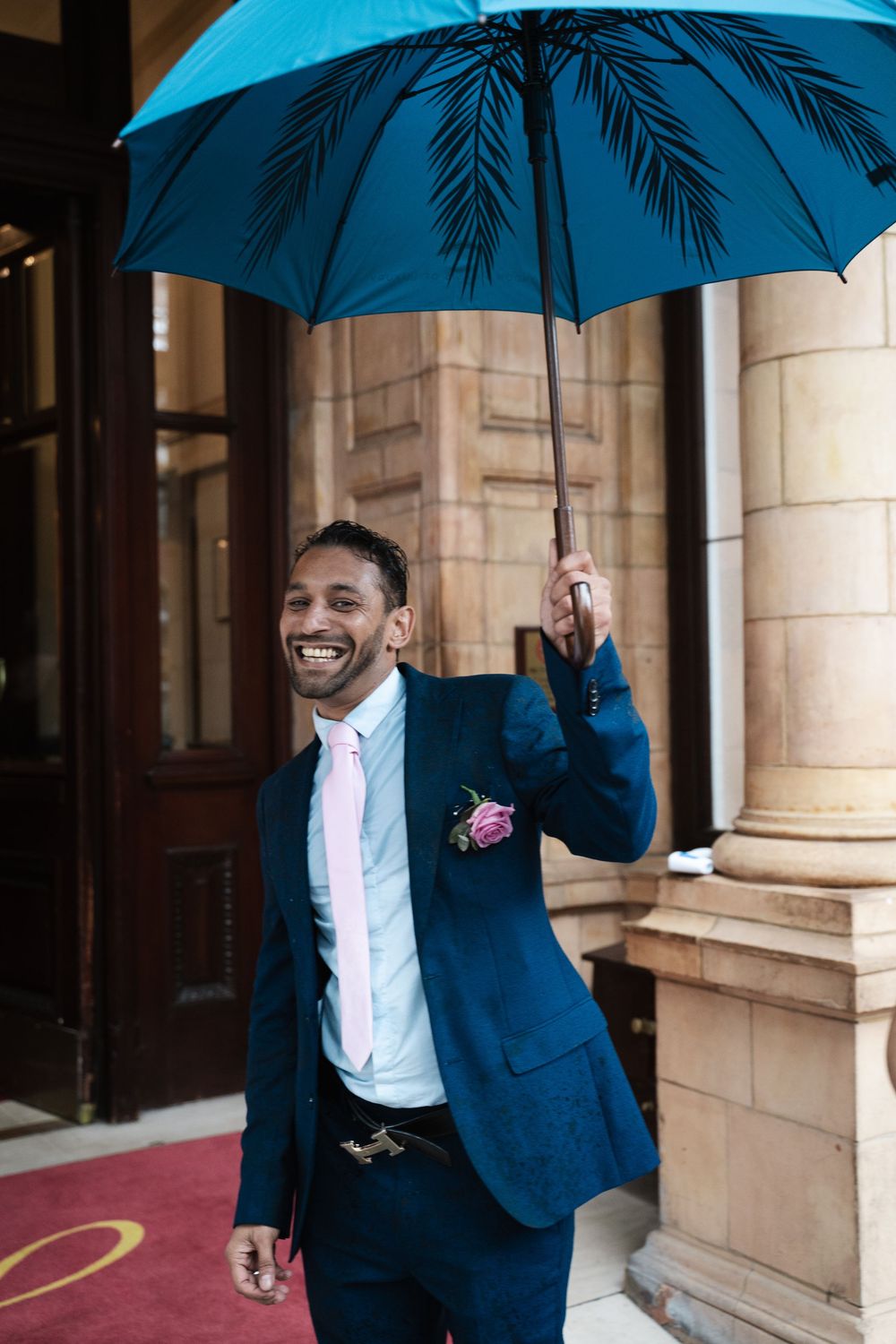 Wedding party member in blue suit holding a bright turquoise umbrella on a rainy day.