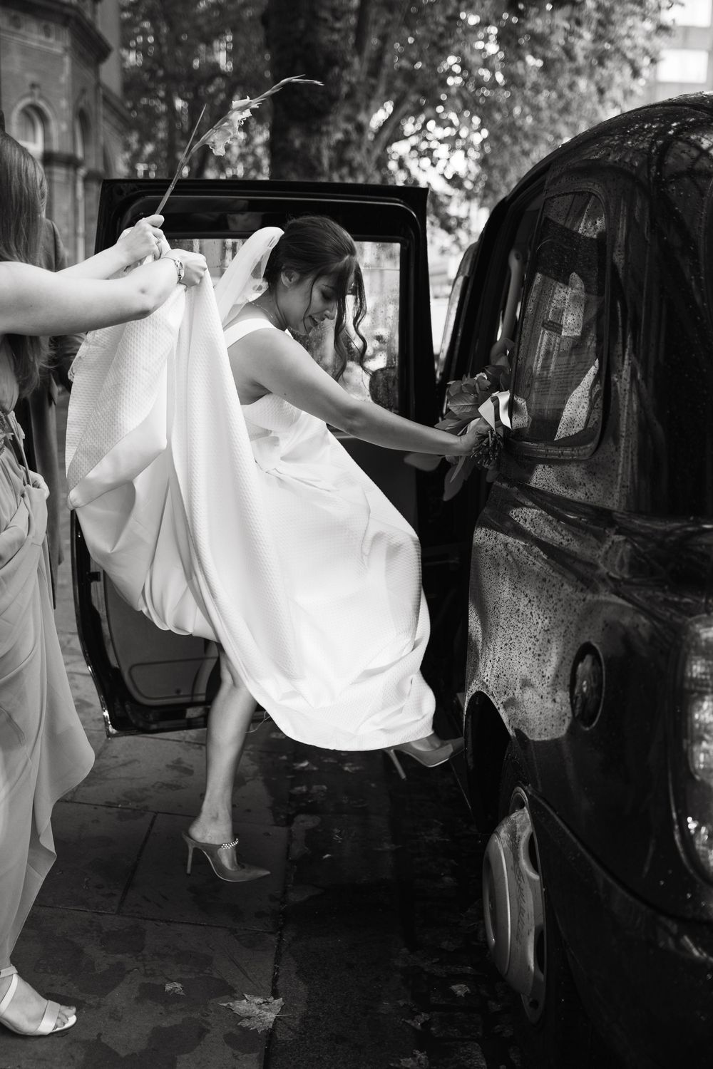 Bride getting into vintage car while holding white wedding dress.