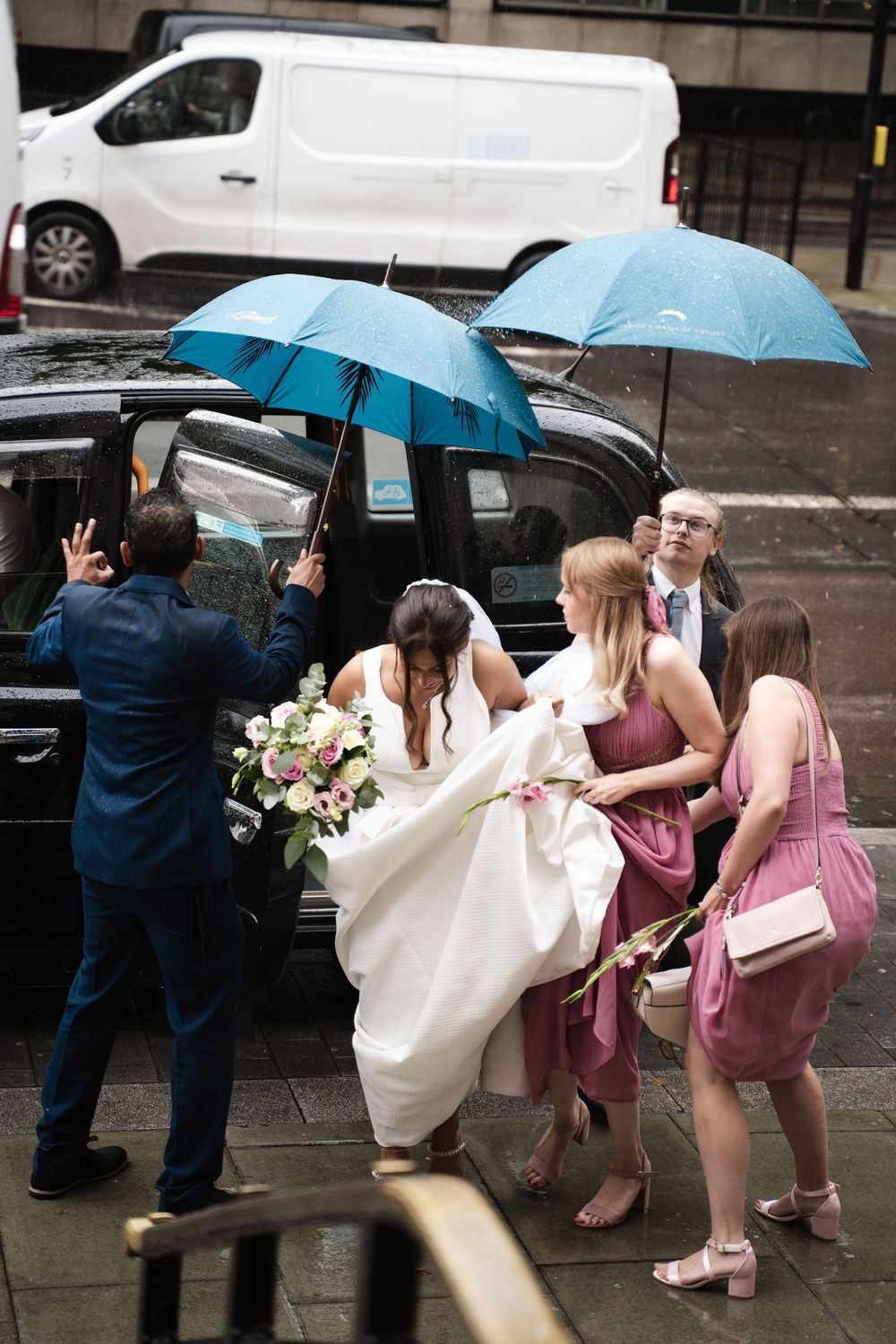 Wedding party helping bride navigate rainy weather with blue umbrellas and pink bridesmaid dresses.