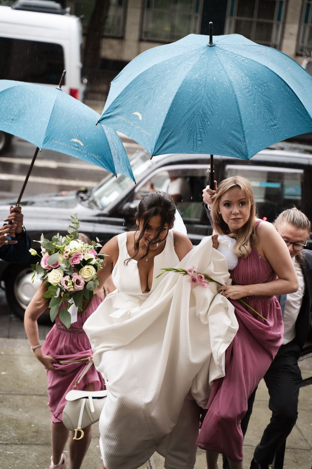 Wedding party hurries through rain under blue umbrellas with bride in white dress and bridesmaids in pink.