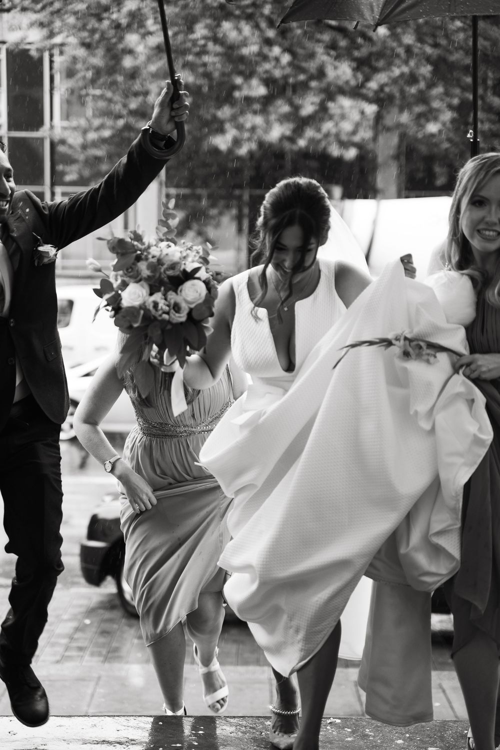 Black and white photo of bridal party rushing through rain with umbrellas and bouquets.