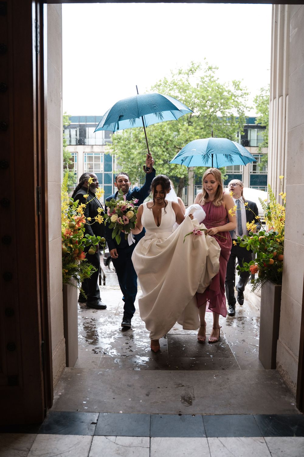 Bride and bridesmaids enter building through doorway while sheltering under bright blue umbrellas.