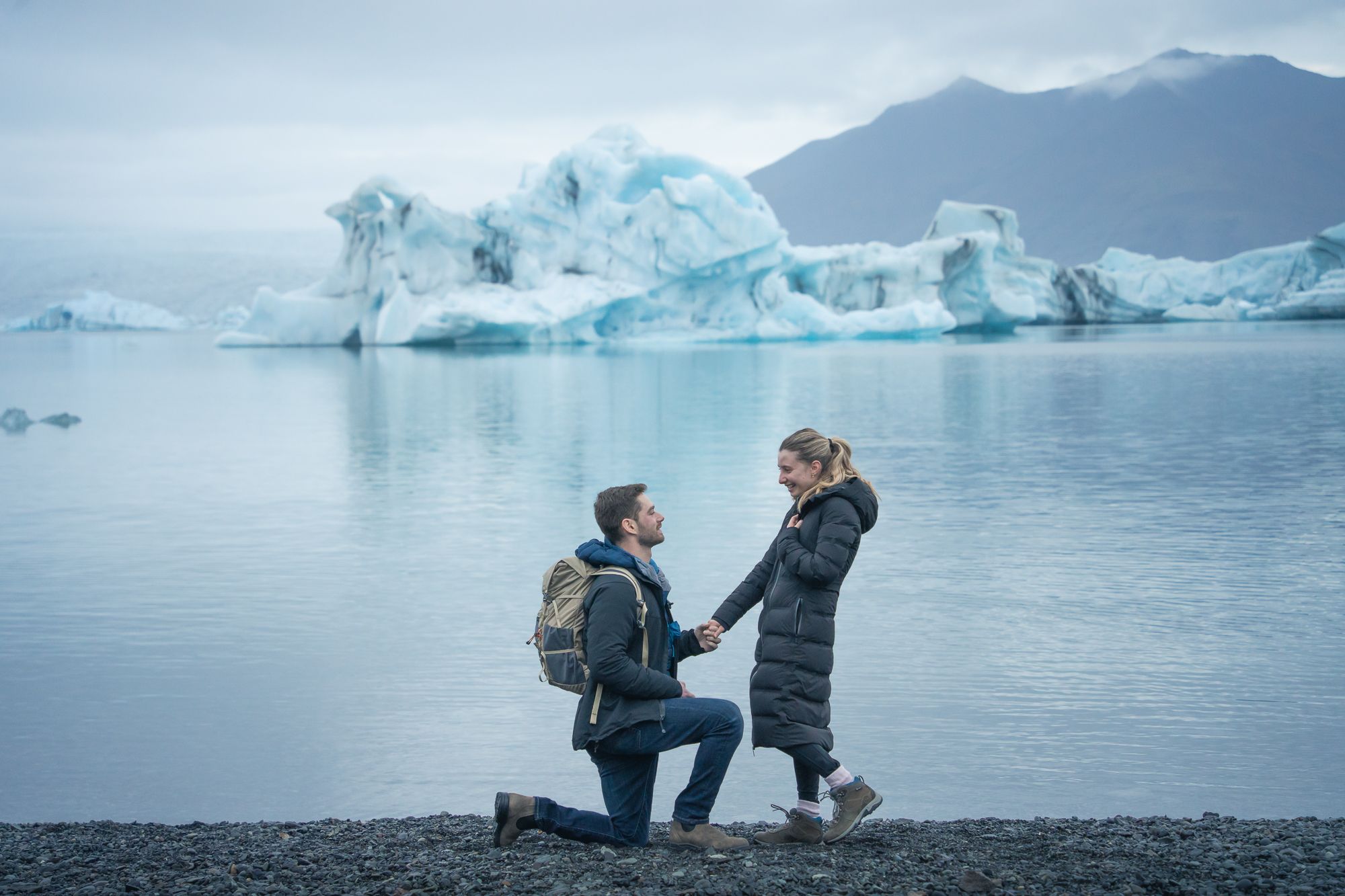Special moment unfolds on black beach with floating icebergs and mountains in background.