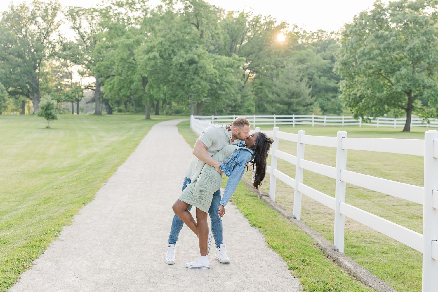 Two people share a playful moment on a gravel path alongside a white fence at a scenic rural property.