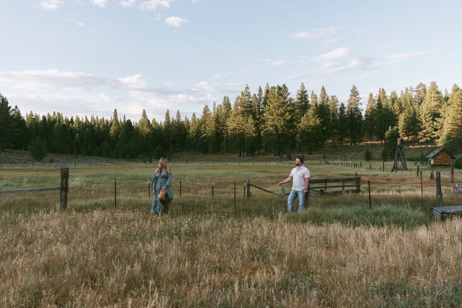 Two people stand in a grassy field with wooden fence and evergreen forest in the background during golden hour in Truckee