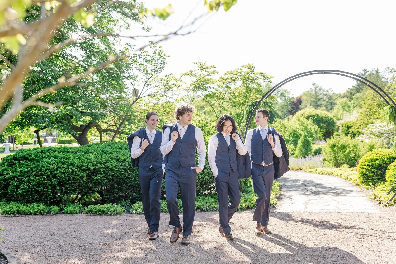 Wedding party members in matching gray vests stroll together along a garden path on a sunny day.