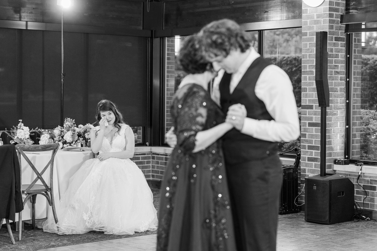 A black and white wedding reception photo showing a couple dancing while a bride sits at a table in the foreground.