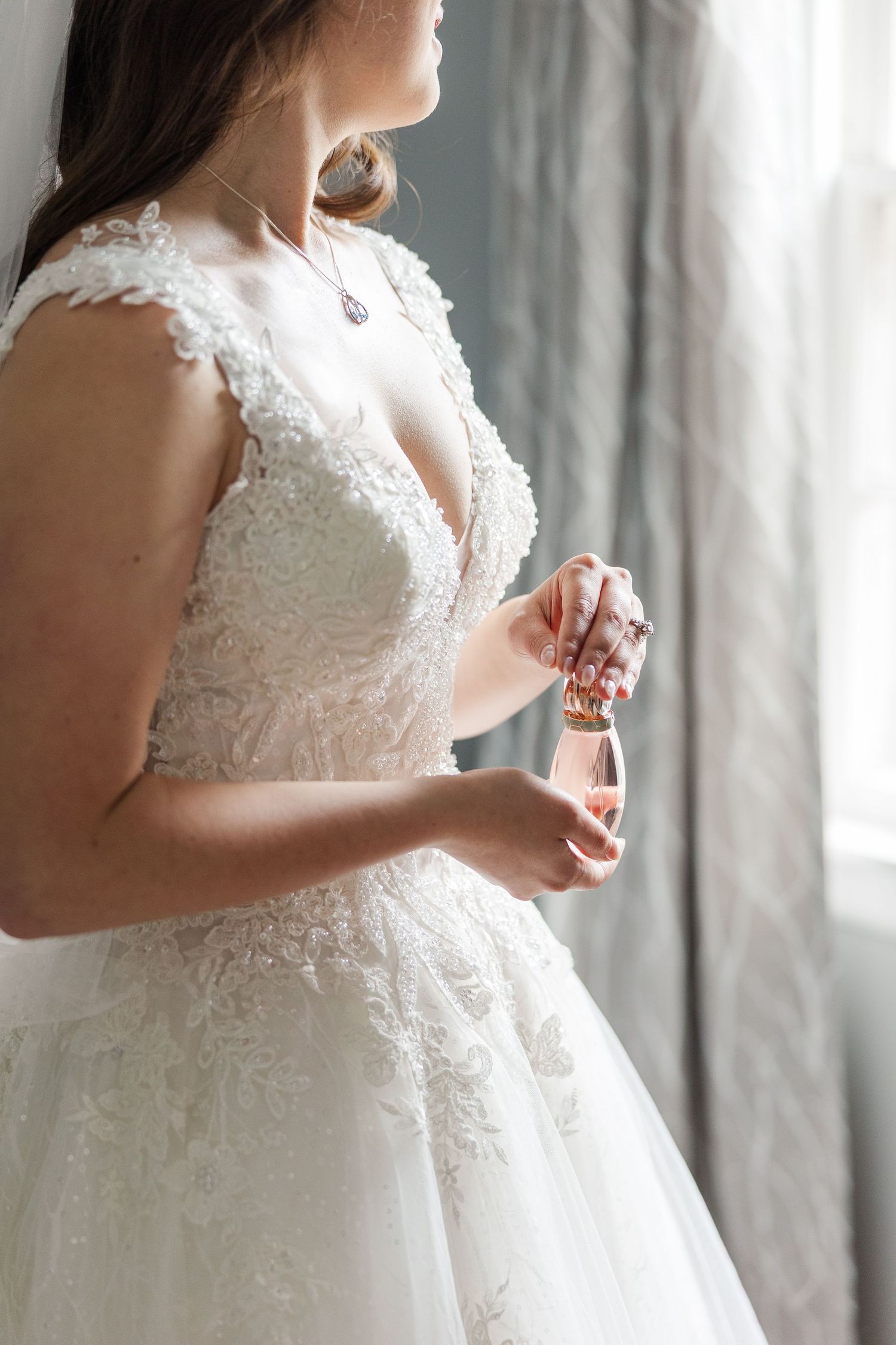 Elegant white lace wedding dress with v-neckline shown in detail as someone holds perfume bottle by window.