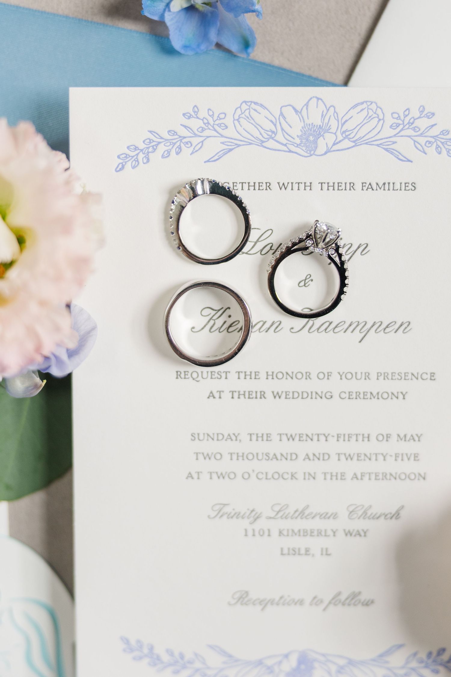 Wedding invitation with delicate blue floral design and three silver wedding rings arranged on white paper.