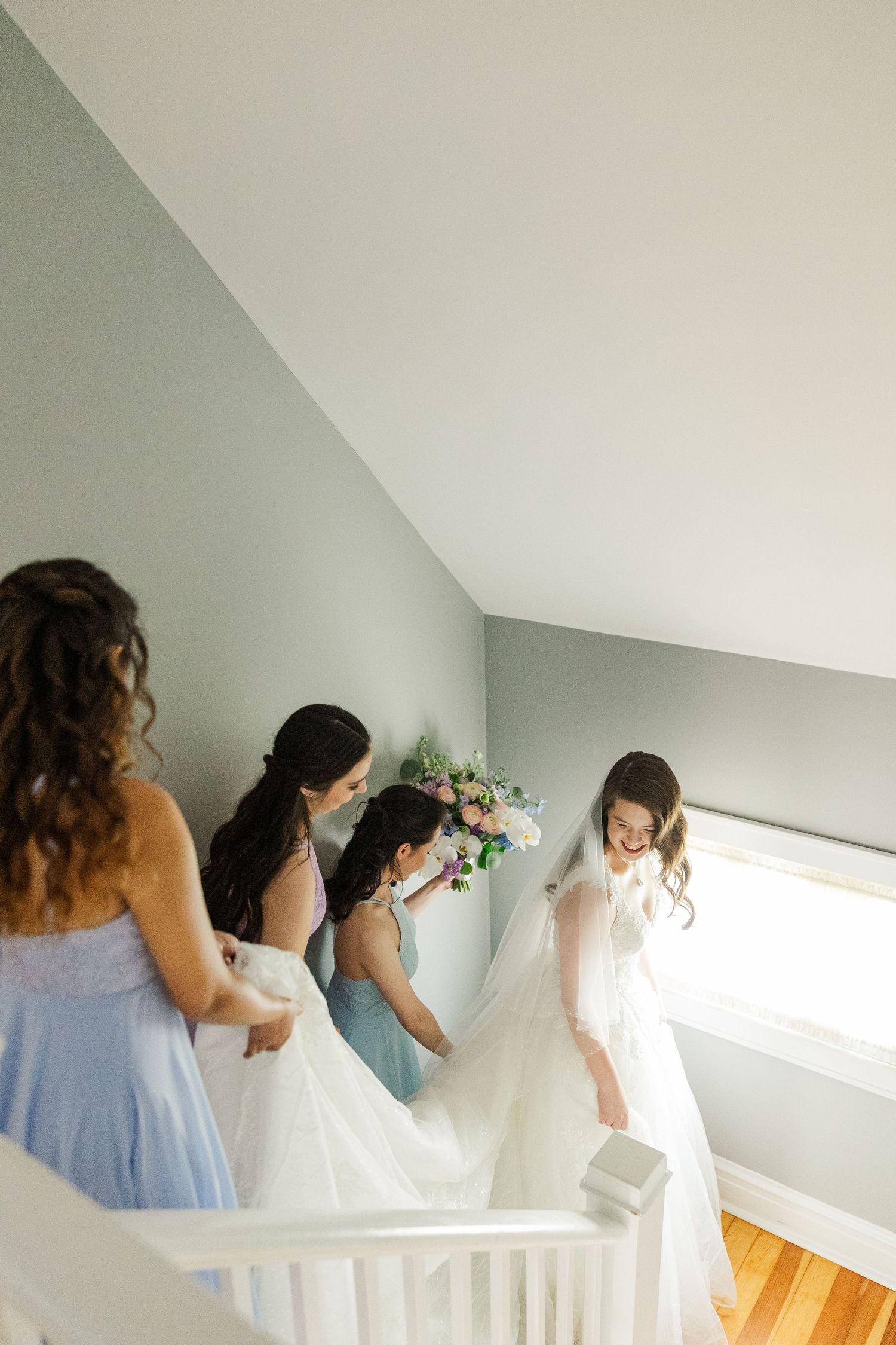 Bridesmaids in light blue dresses help adjust a bride's gown while ascending a bright white staircase.