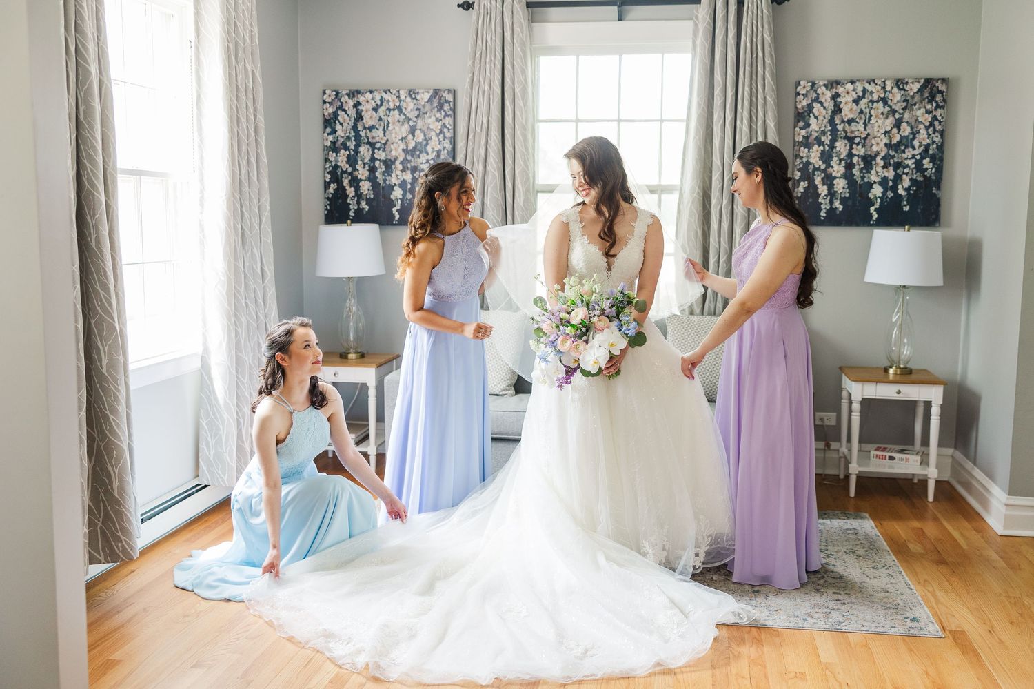 A bridal party gathers in an elegant room with black and white floral artwork, pastel dresses, and hardwood floors.