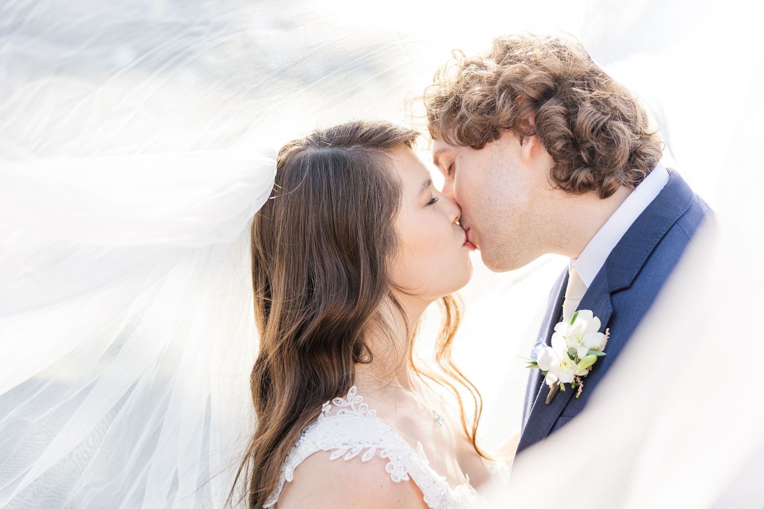 A romantic couple in wedding attire share a kiss under a flowing white veil in an elegant outdoor setting.