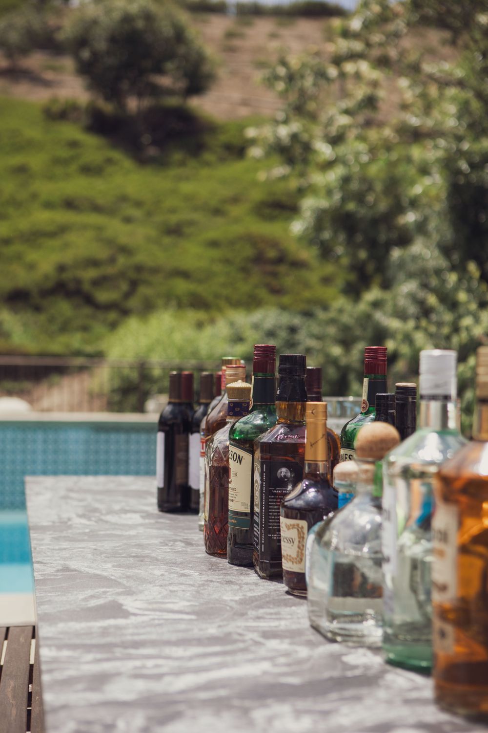 Various liquor and wine bottles lined up on a stone ledge near a swimming pool with hills in the background.