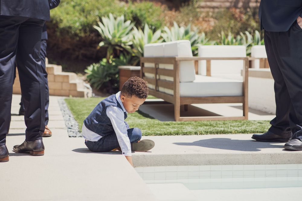 A small child in a navy shirt sits on the ground between two adults standing on a patio with outdoor furniture.