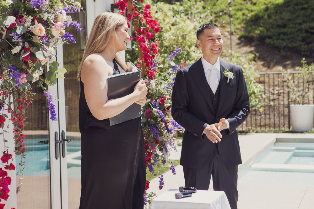 Wedding officiant in black dress stands at flower-adorned altar with groom in tuxedo during outdoor poolside ceremony.