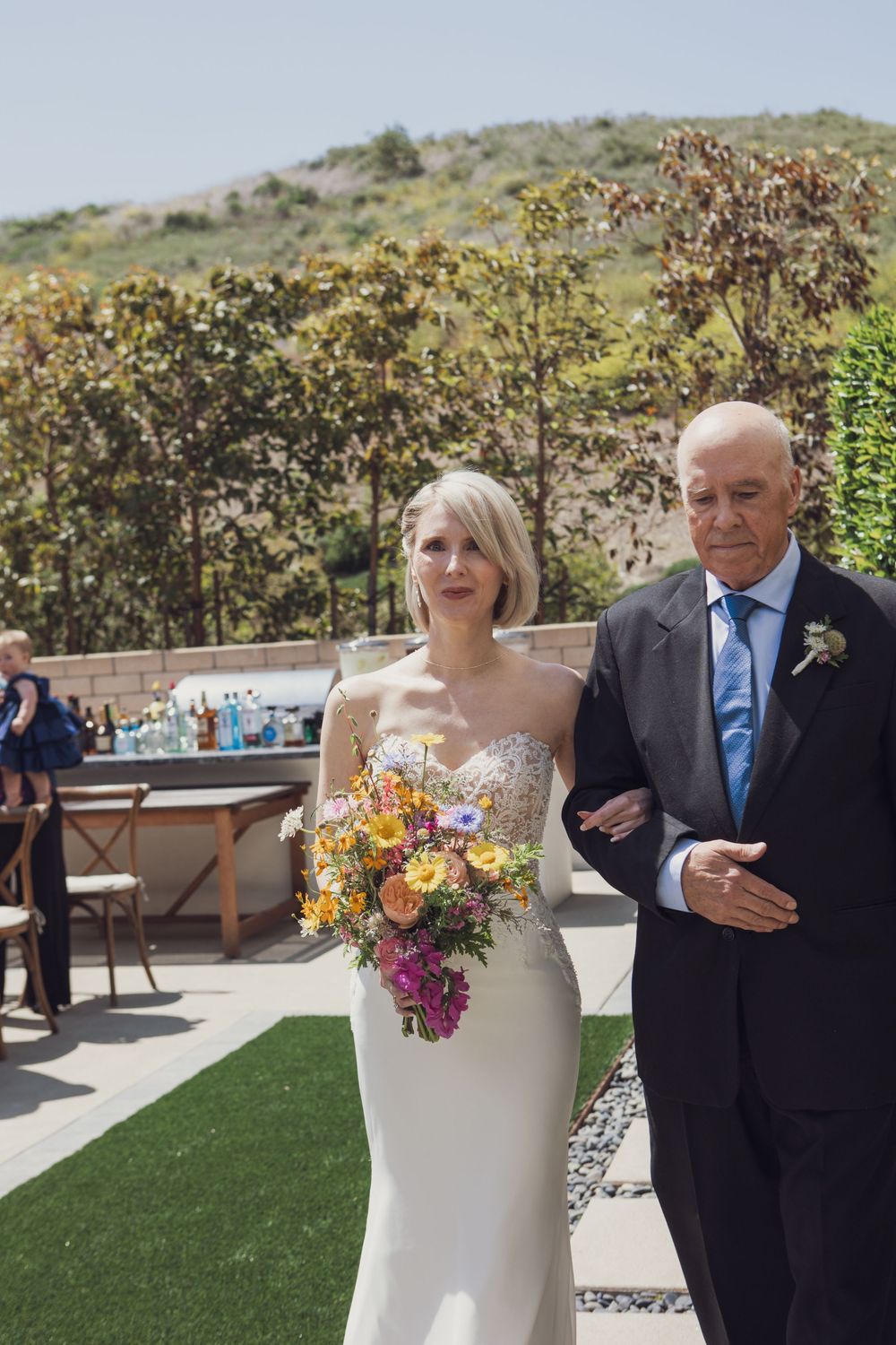 Two people at an outdoor wedding ceremony walking together on a paved area with greenery in the background.