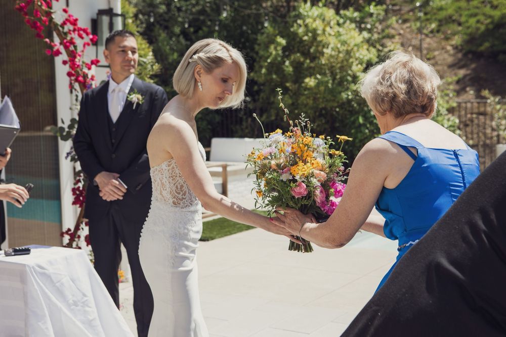 Someone in a sleek white wedding dress receives a beautiful orange and yellow bouquet during an outdoor ceremony.