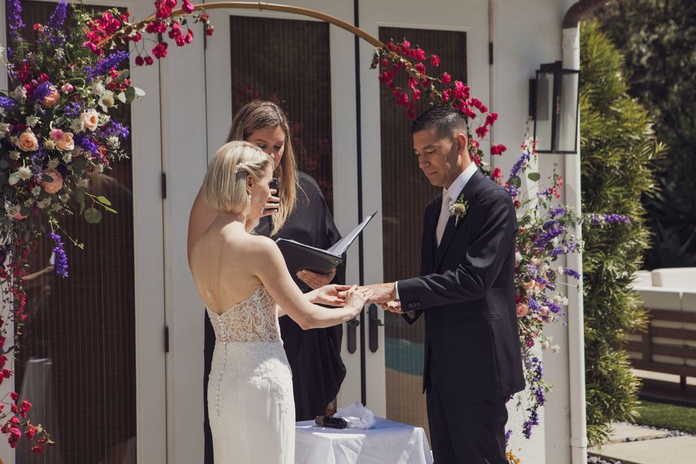 A romantic outdoor wedding ceremony takes place beneath an arbor adorned with vibrant pink and purple blooming flowers.