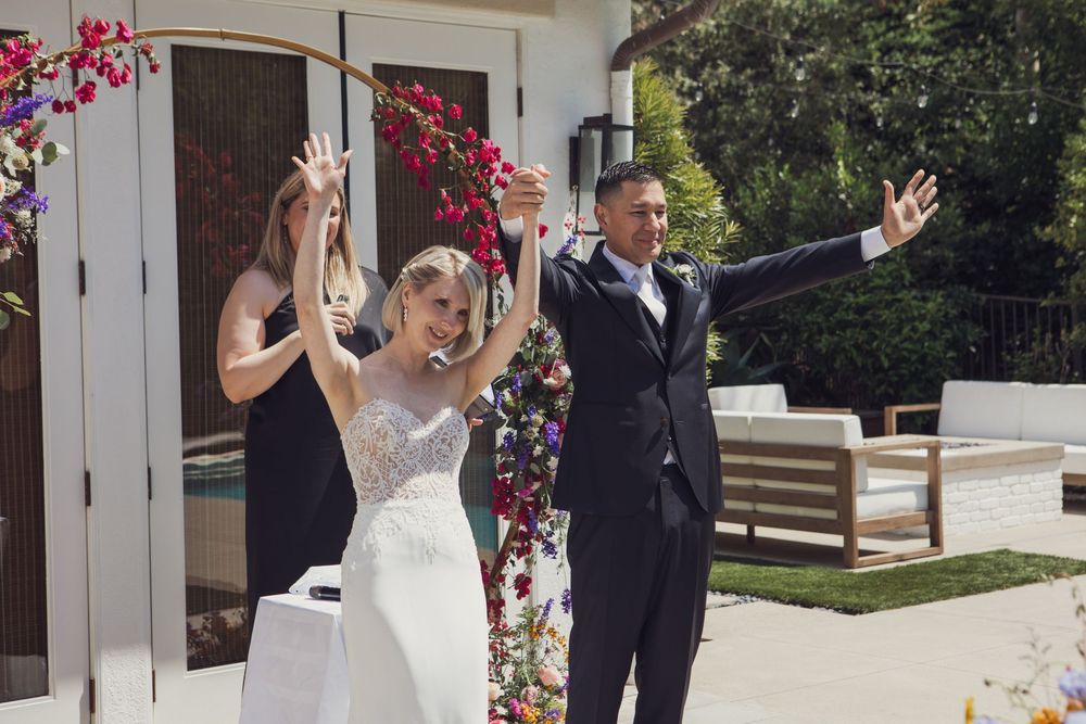 A newlywed couple celebrates with raised arms under a pink flower archway on their wedding day at an outdoor venue.