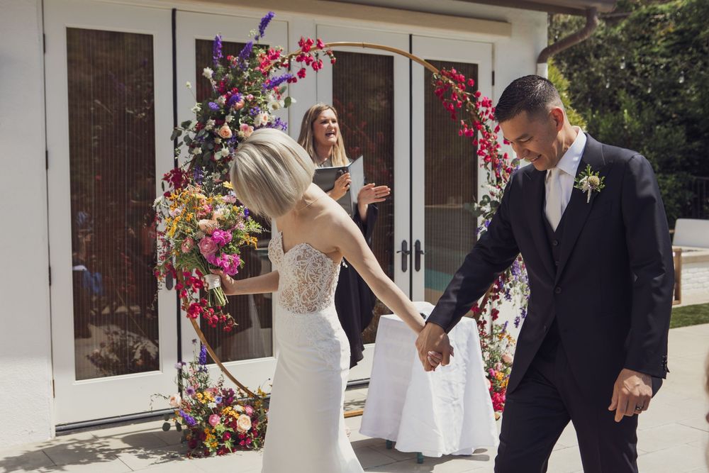 A couple joyfully exits their ceremony under a floral arch while holding hands in a bright outdoor setting.