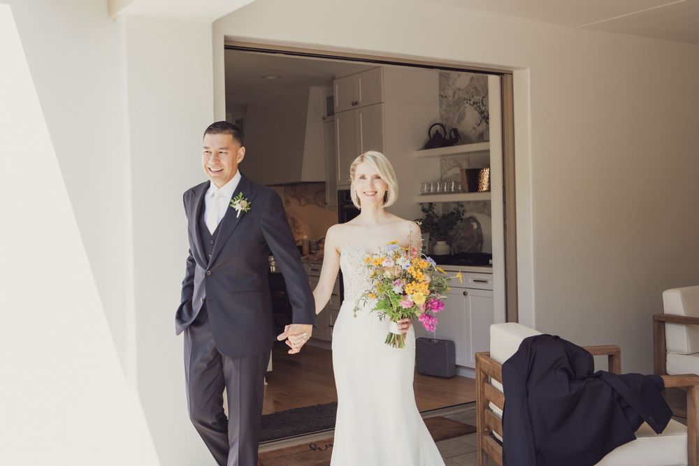 Newlywed couple exits doorway together in wedding attire with colorful bouquet during sunny celebration.