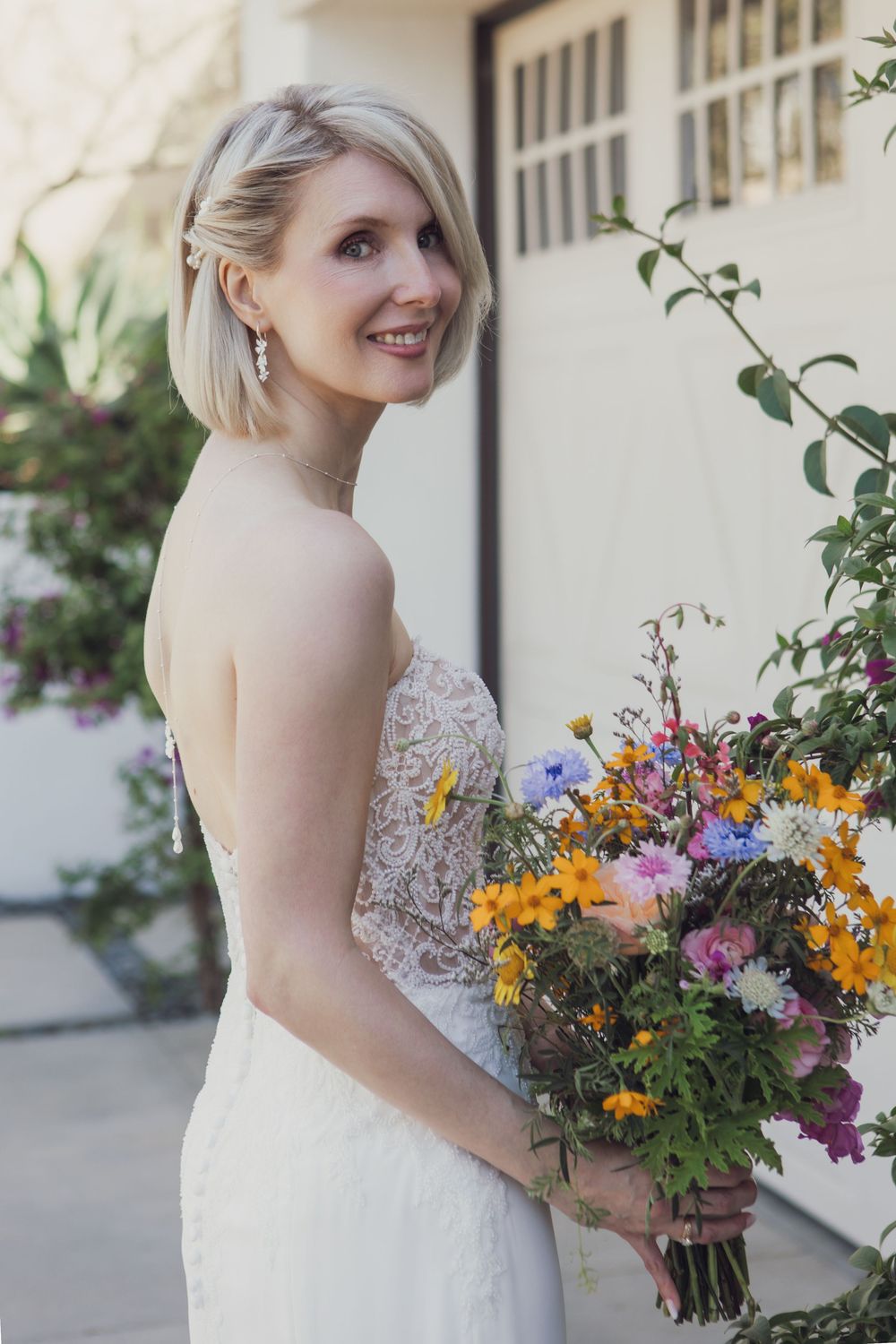 Beautiful bride in white lace dress holds colorful wildflower bouquet with yellow and purple blooms against light wall.