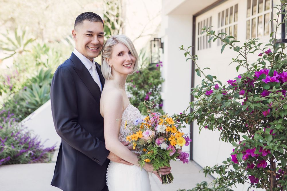 A couple in wedding attire stands together outdoors by flowering plants with the bride holding a colorful bouquet.