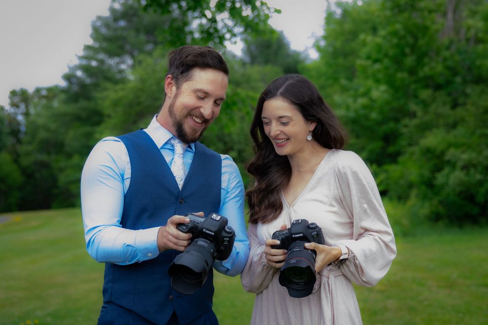 Two photographers review photos on their DSLR cameras while standing together outdoors on a green lawn.