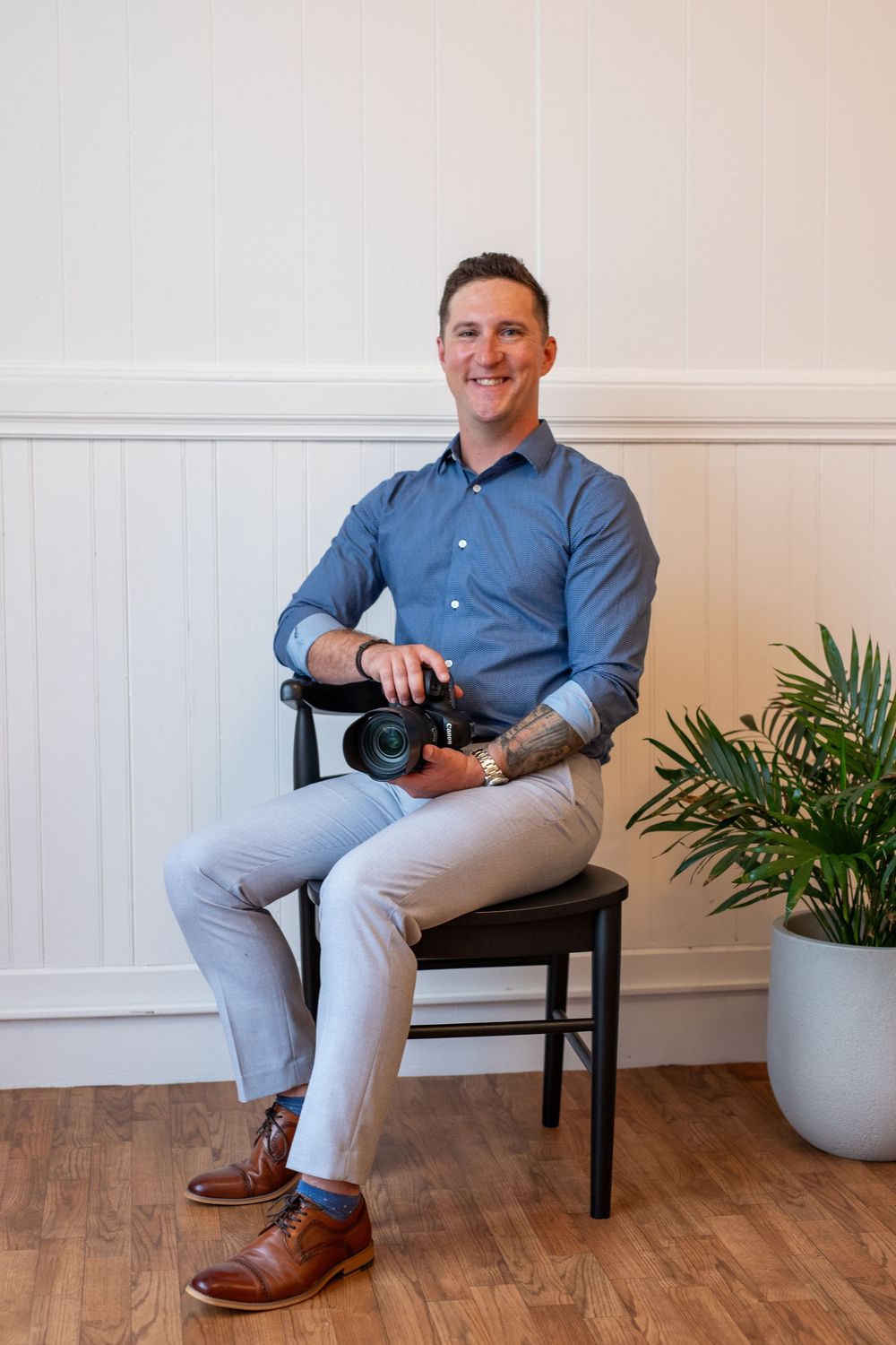 Person in blue dress shirt and light gray pants sitting on dark wooden bench against white wall with potted plant.