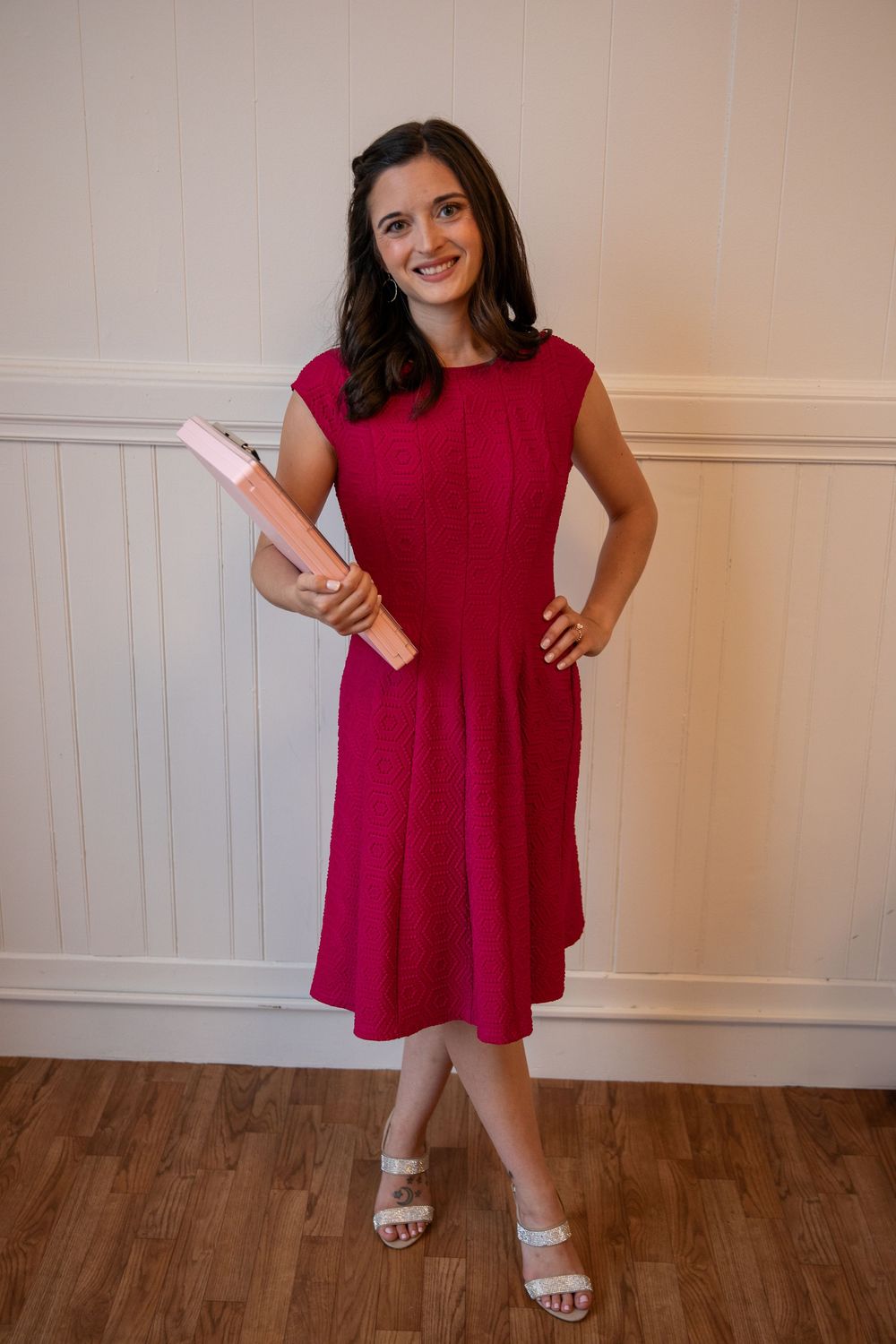 Someone wearing a sleeveless red midi dress with sandals poses holding a pink diploma holder against a white wall.