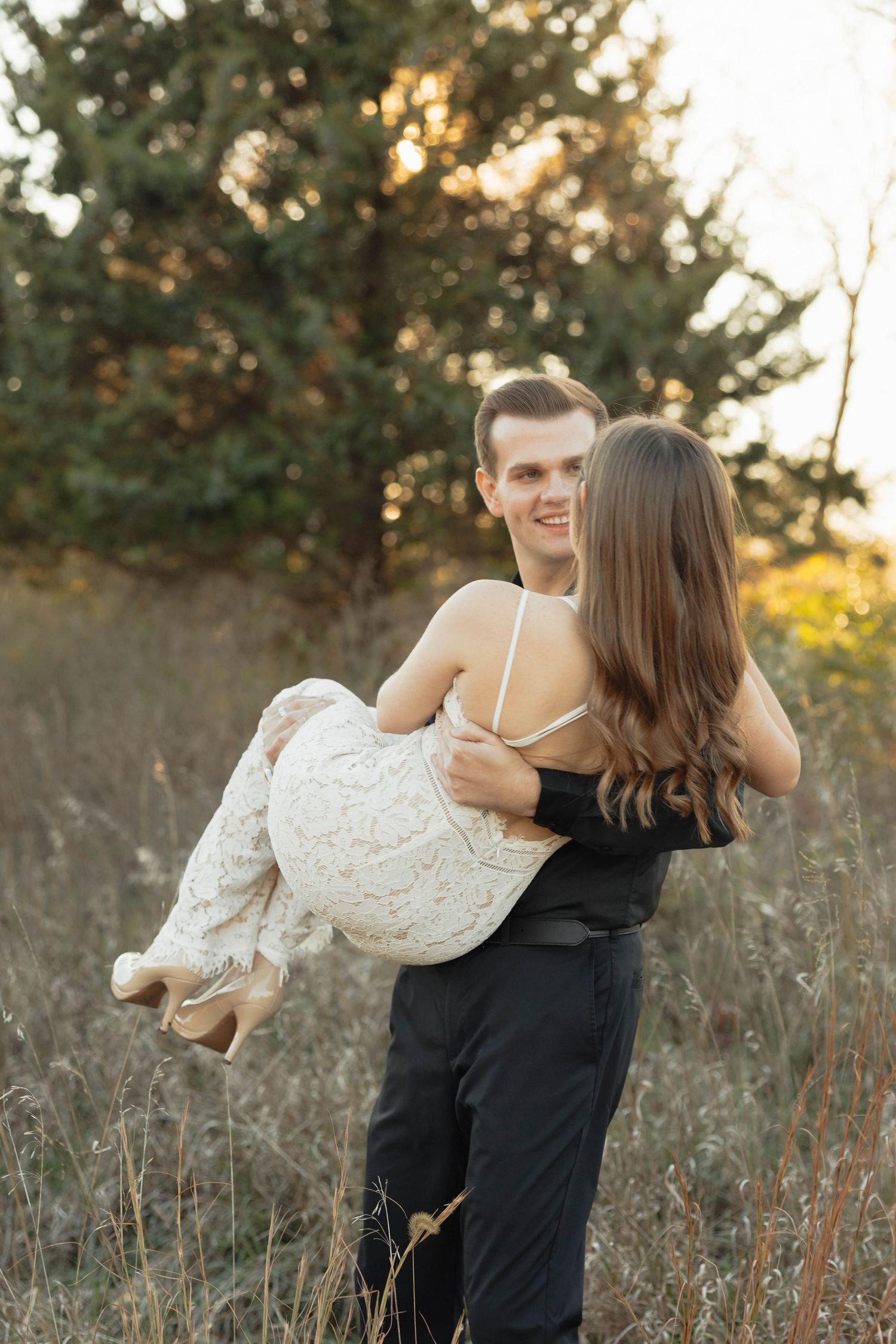 A couple embraces playfully in a golden field at sunset, with the partner in black lifting and spinning their companion.