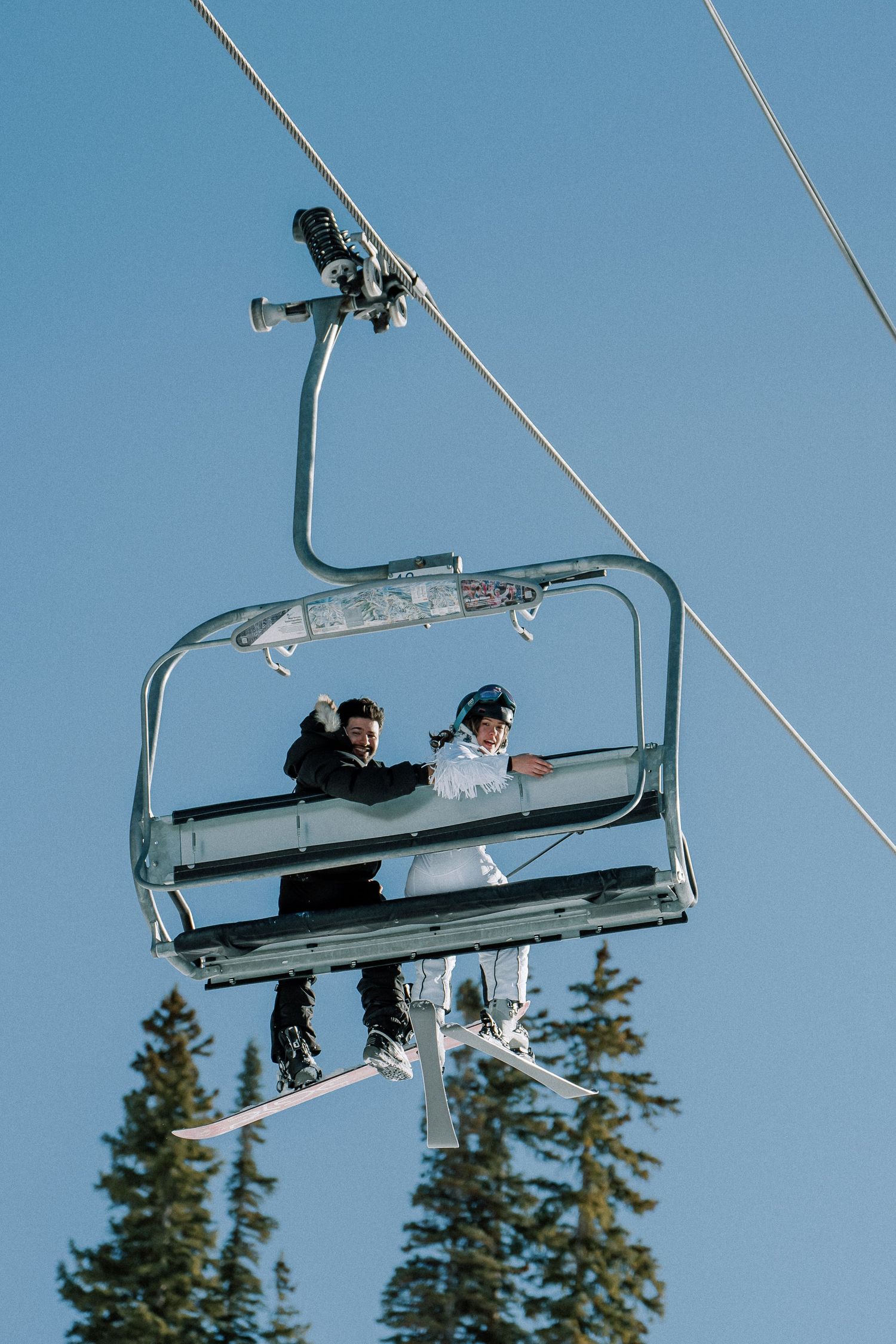 Two skiers ride a chairlift against a blue sky with evergreen trees visible in the background at a ski resort.