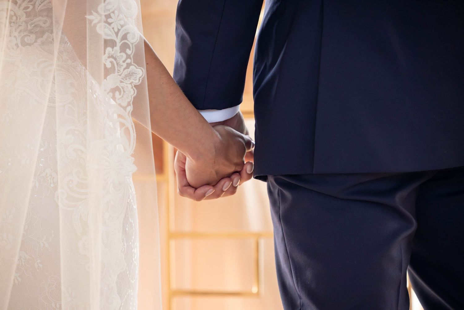 Close-up view of a couple holding hands during a wedding ceremony with white veil and navy suit visible.